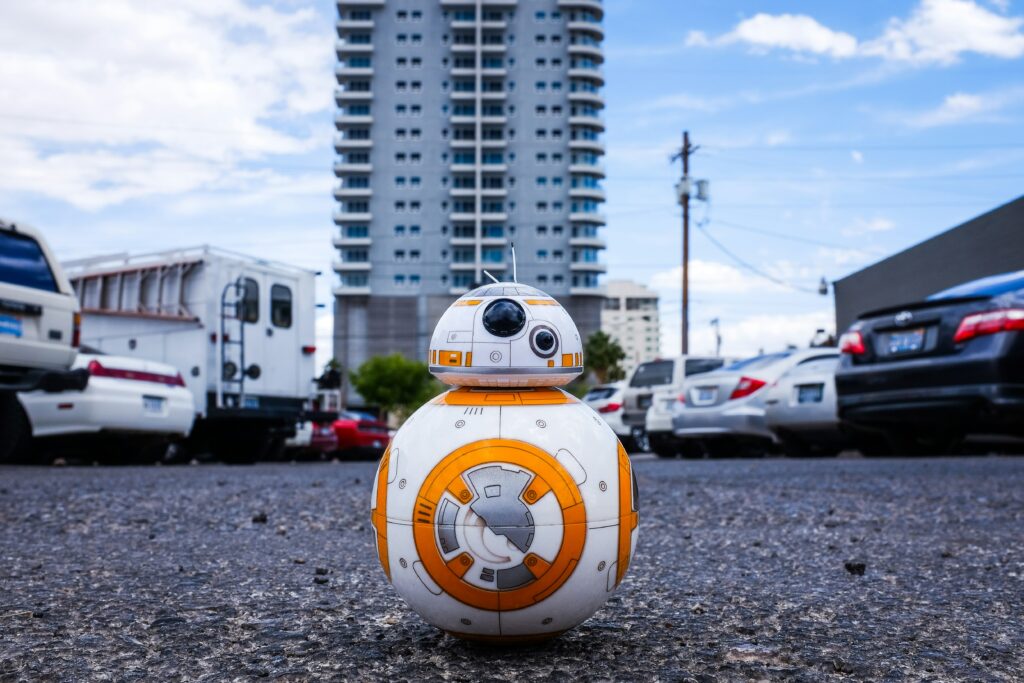 A BB-8 robot toy on a parking lot with a tall building in the background.