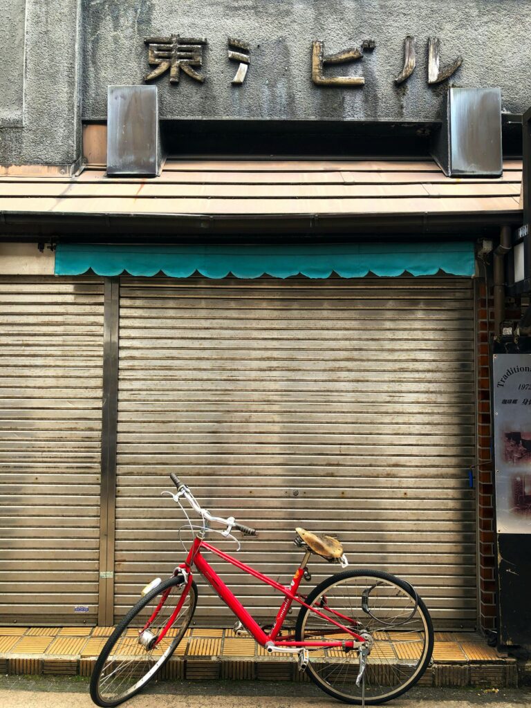 A red bicycle leans against a closed storefront with faded signs and a rolled metal shutter.